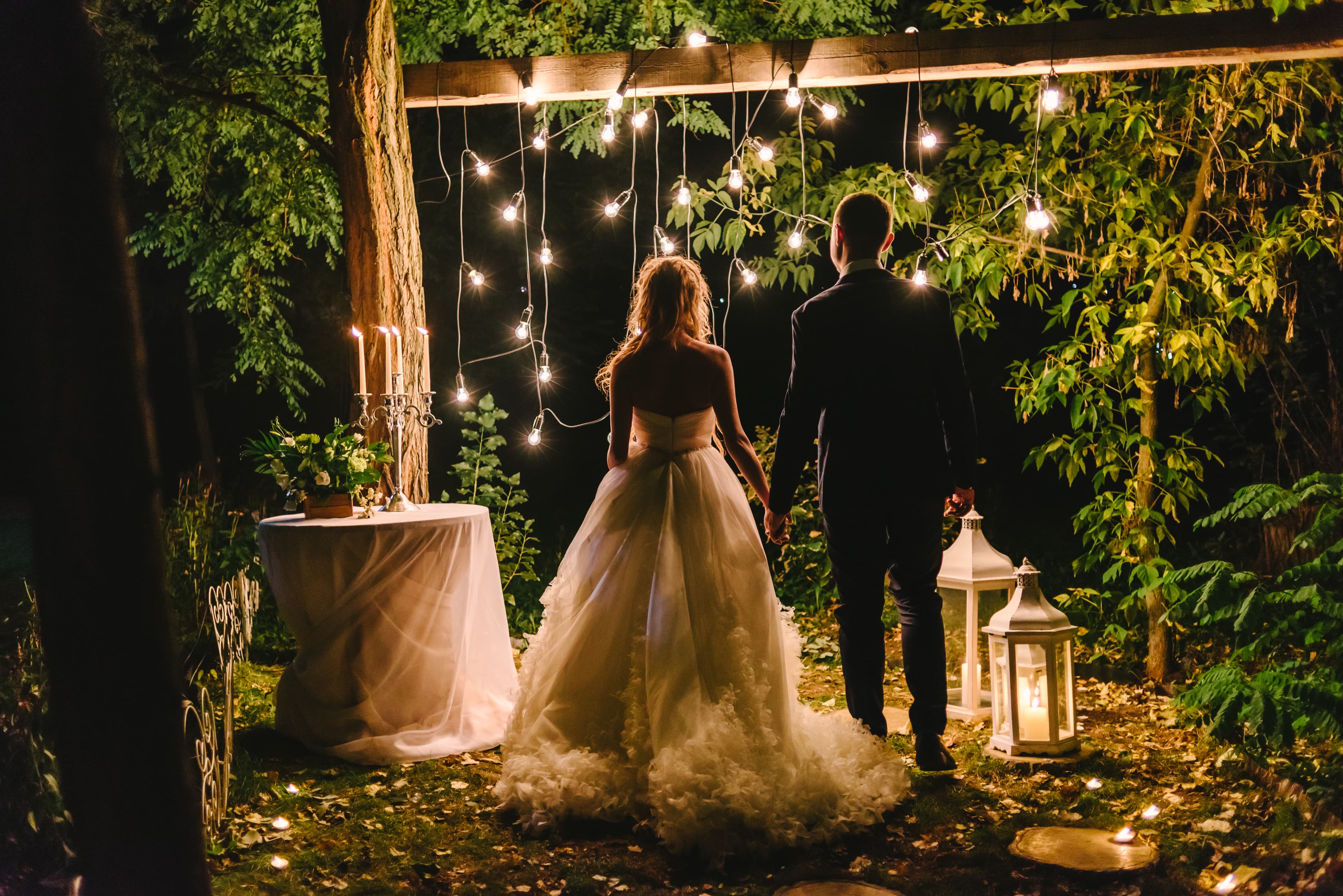 bride and groom standing together holding hands at night in front of string lights