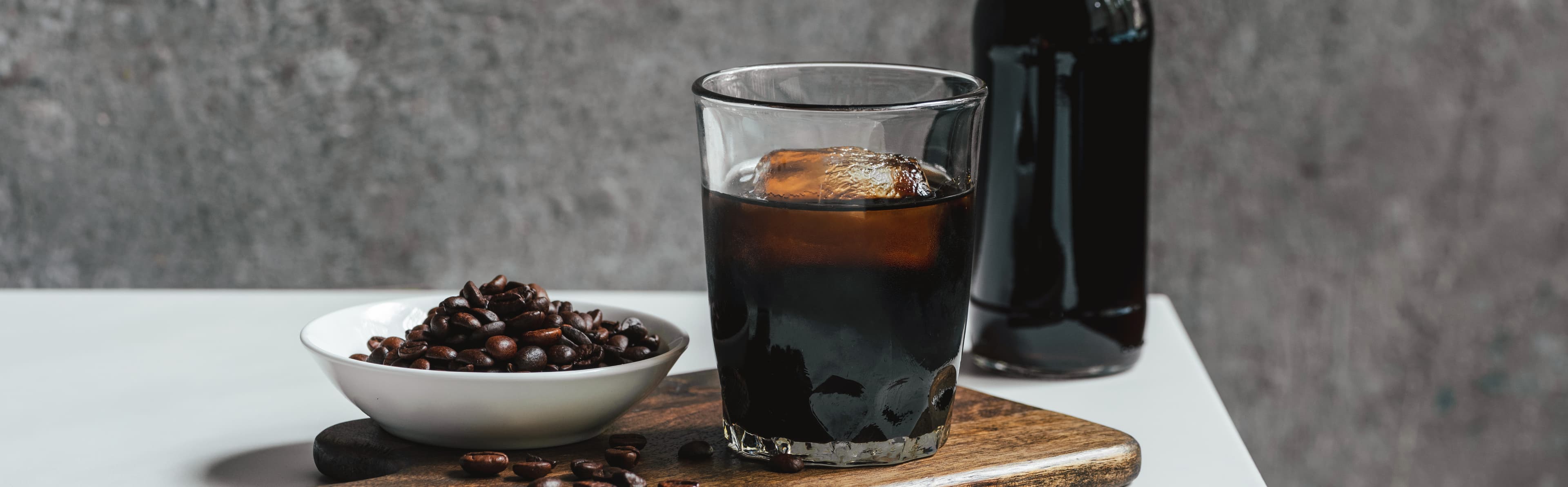 Grey Background with White Table in the Foreground holding a wooden cutting board with a white bowl of dark roasted coffee beans next to a glass of cold brew coffee with one large cube of ice