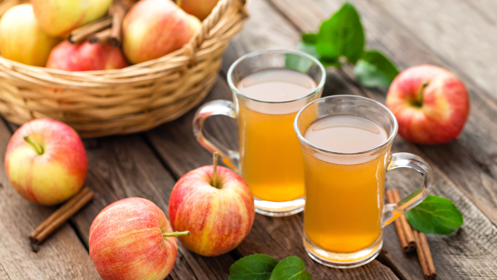 What is Apple Cider | two glasses of apple cider on a pic-nic table next to a woven basket of honeycrisp apples. there are a few apples on the table alongside cinnamon sticks and apple leaves