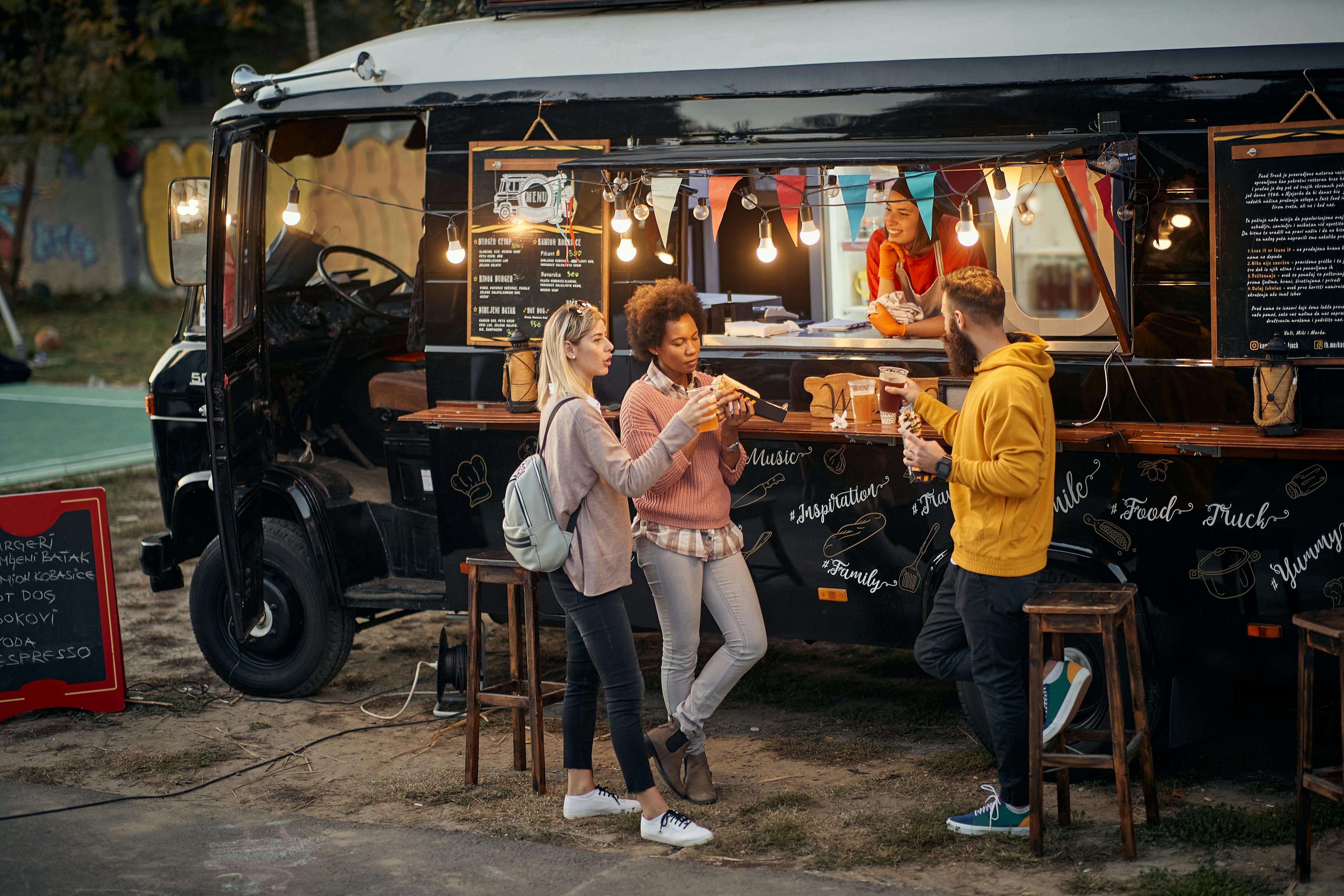two women and a man stand in front of a black food truck enjoying food and drink served by a woman in a red shirt. the food truck has a menu on the side 