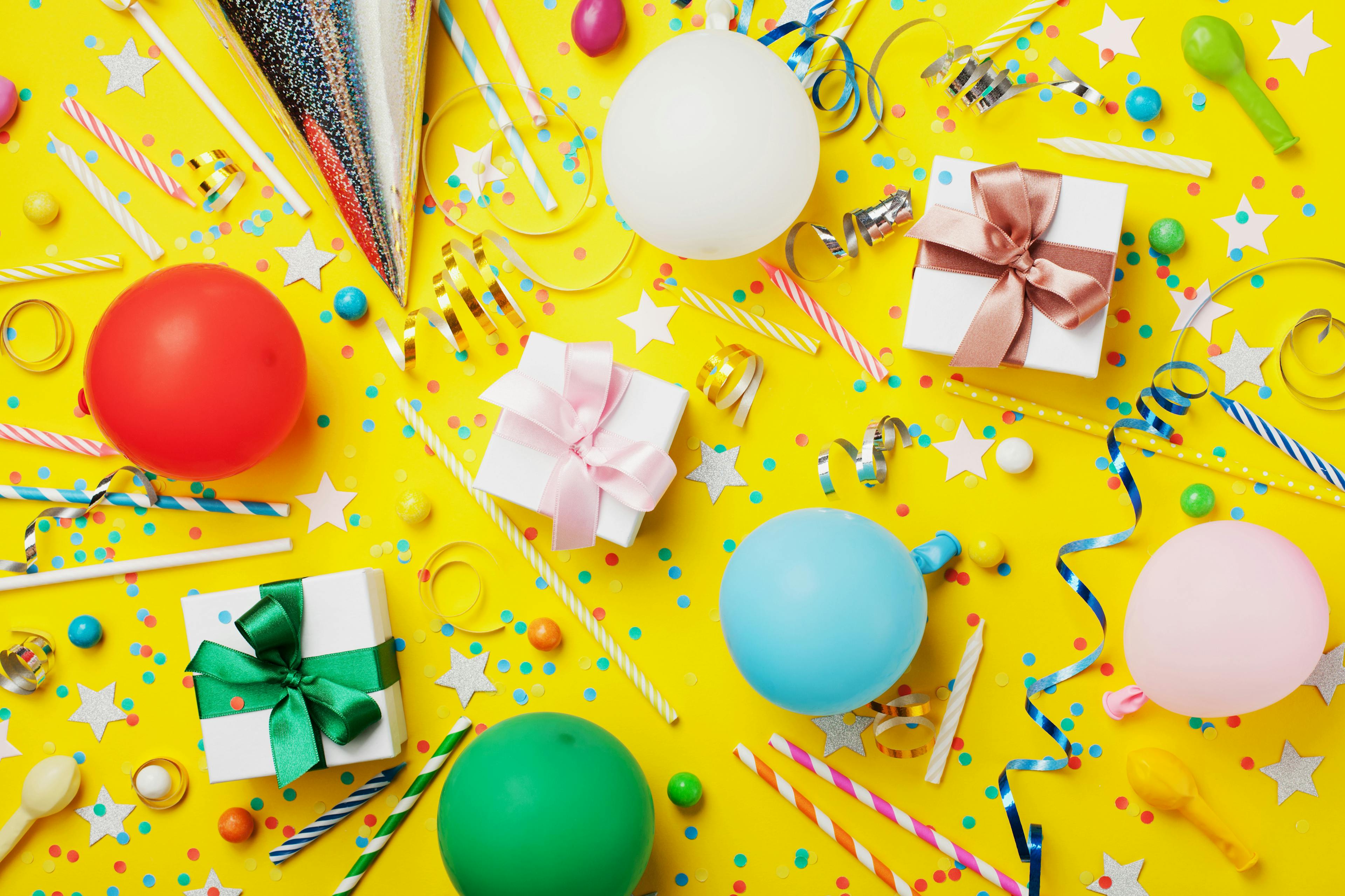 Yellow Background covered in small multi-colored dot confetti. Three small packages wrapped in bows and a party hat