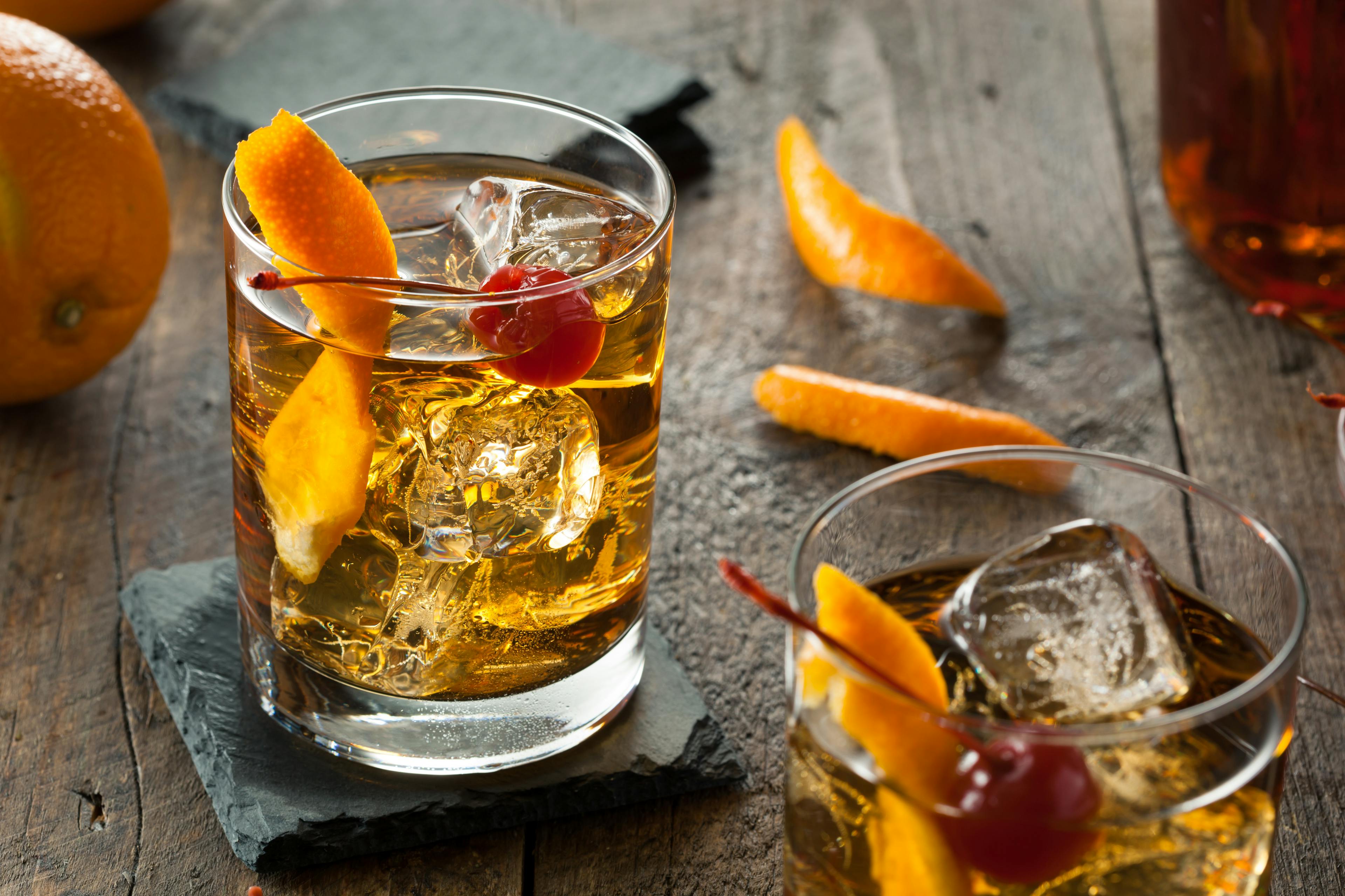 This image shows an old fashioned cocktail with a twist of orange and a cherry sitting in a short heavy based glass cocktail glass that sits on a black slate coaster all sitting on a wooden table