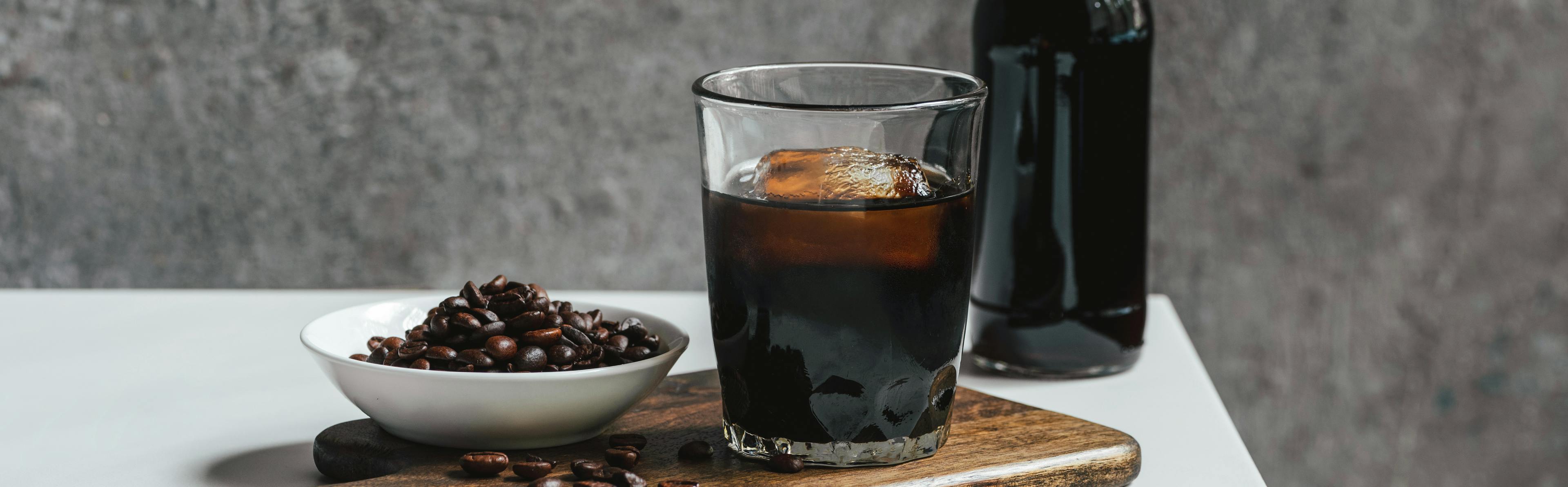 Grey Background with White Table in the Foreground holding a wooden cutting board with a white bowl of dark roasted coffee beans next to a glass of cold brew coffee with one large cube of ice