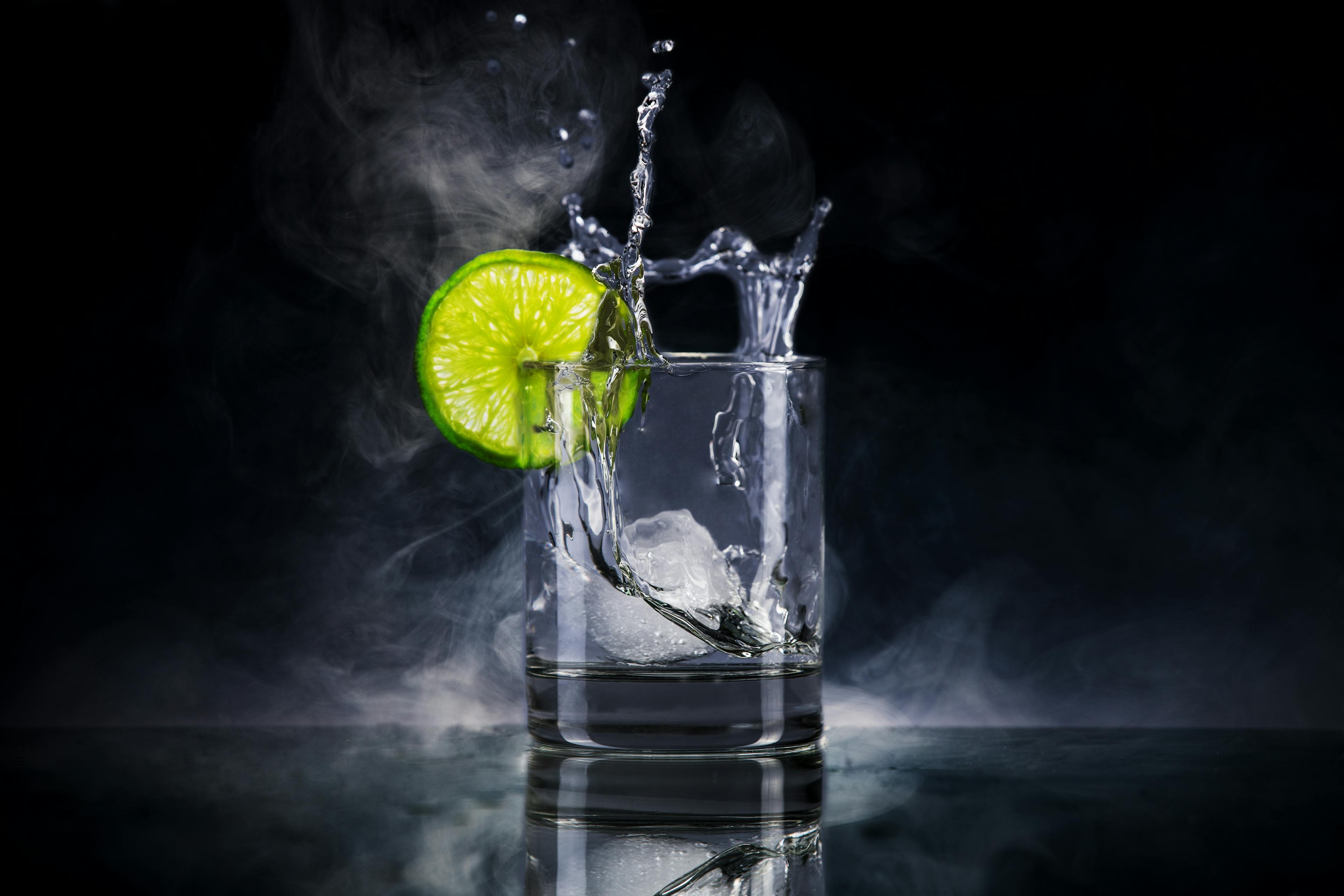 clear low ball glass holds vodka and the action shows a cube of ice being dropped into the glass, which has a slice of lime on the rim with a smoky black background